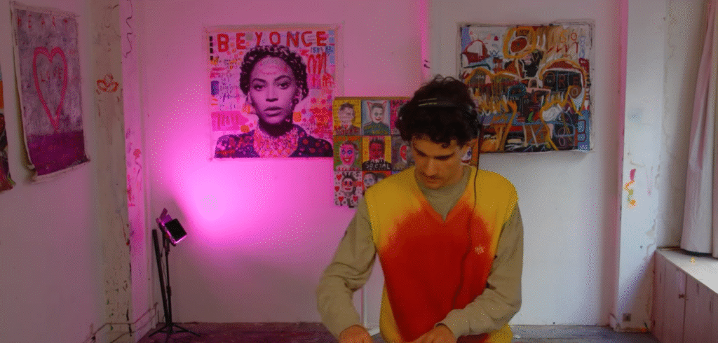 Sur cette photo, Paradise777, DJ et producteur français, est concentré sur ses platines dans un espace artistique atypique. Le décor coloré mêle street art et lumière rose, reflétant parfaitement l’univers hybride et créatif de cet artiste qui fusionne techno mentale, breaks et drum & bass. On aperçoit au mur une grande toile représentant Beyoncé, ajoutant une touche d’inspiration pop à l’ambiance électrique du lieu.