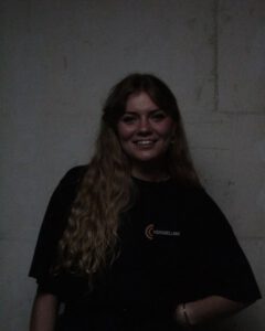 Portrait d'une jeune femme aux longs cheveux blonds ondulés, souriante, portant un t-shirt noir avec le logo 'VERSNELLING' sur la poitrine, posant devant un mur clair dans une ambiance sombre.