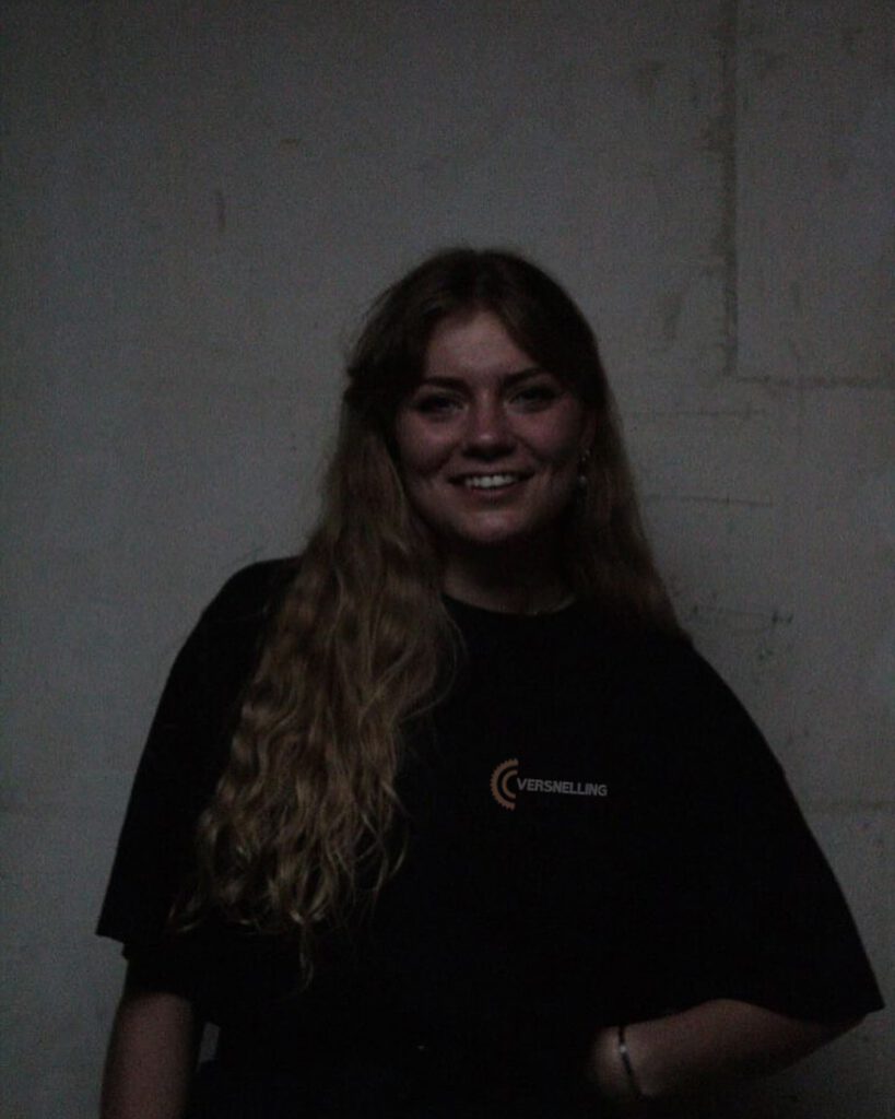 Portrait d'une jeune femme aux longs cheveux blonds ondulés, souriante, portant un t-shirt noir avec le logo 'VERSNELLING' sur la poitrine, posant devant un mur clair dans une ambiance sombre.