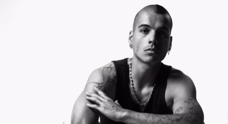 Sur une photo en noir et blanc, un homme est assis sur une chaise, vêtu d'un débardeur noir et d'une chaîne métallique autour du cou. Il arbore plusieurs tatouages sur les bras et porte une boucle d'oreille visible. L'homme pose de façon étendue, un avant-bras appuyé sur l'autre. L'image est prise sur un fond blanc épuré, mettant en valeur son allure et ses tatouages.