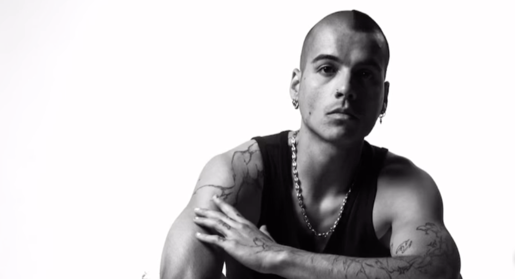 Sur une photo en noir et blanc, un homme est assis sur une chaise, vêtu d'un débardeur noir et d'une chaîne métallique autour du cou. Il arbore plusieurs tatouages sur les bras et porte une boucle d'oreille visible. L'homme pose de façon étendue, un avant-bras appuyé sur l'autre. L'image est prise sur un fond blanc épuré, mettant en valeur son allure et ses tatouages.