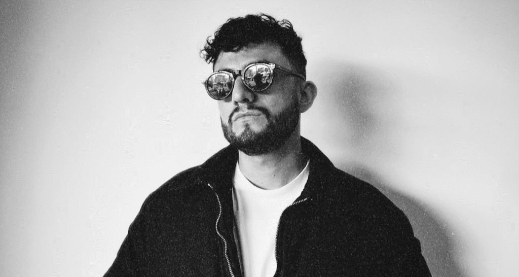 Portrait en noir et blanc d'un homme à la chevelure bouclée et aux lunettes, portant une veste foncée sur un t-shirt clair, photographié devant un mur uni.