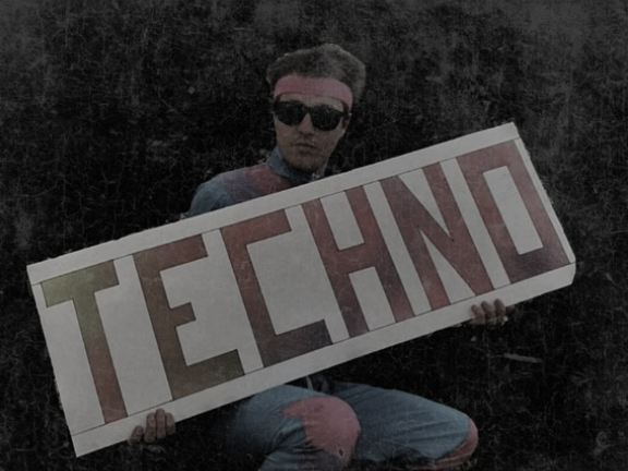 Homme assis portant des lunettes noires et un bandeau rose, tenant un grand panneau blanc avec le mot « TECHNO » inscrit en lettres capitales rouges et vertes, sur un fond sombre à l'effet vieilli.